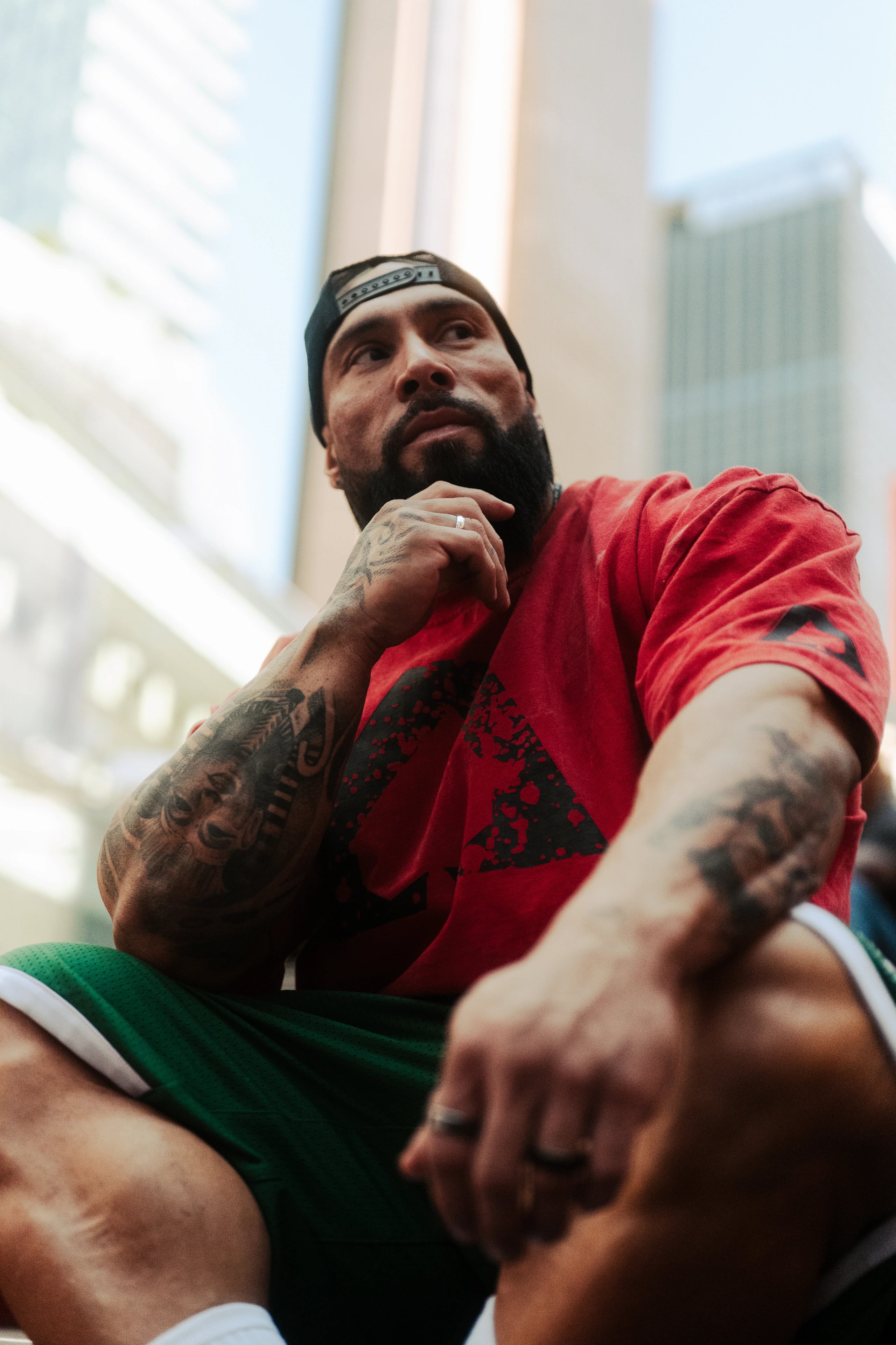 A tattooed person in a red shirt sits among city buildings.