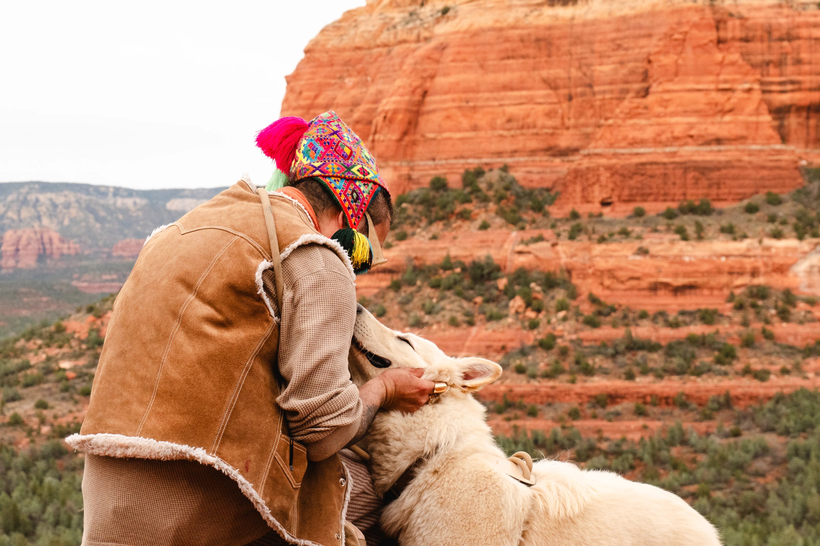 A rider in a colorful knit hat sits with a light-colored horse before red rock cliffs.