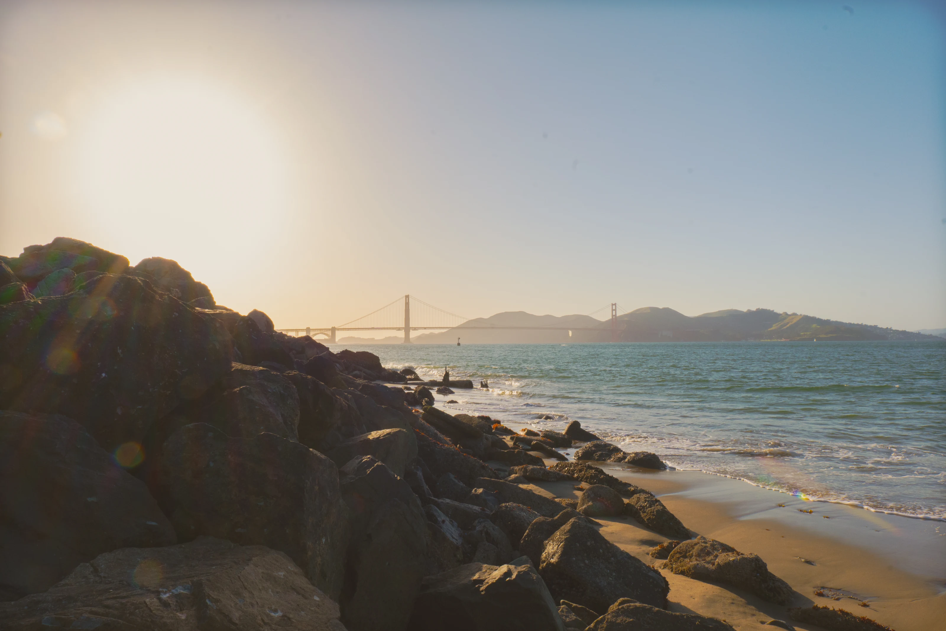 The Golden Gate Bridge crosses San Francisco Bay beyond a rocky shoreline in warm evening light.