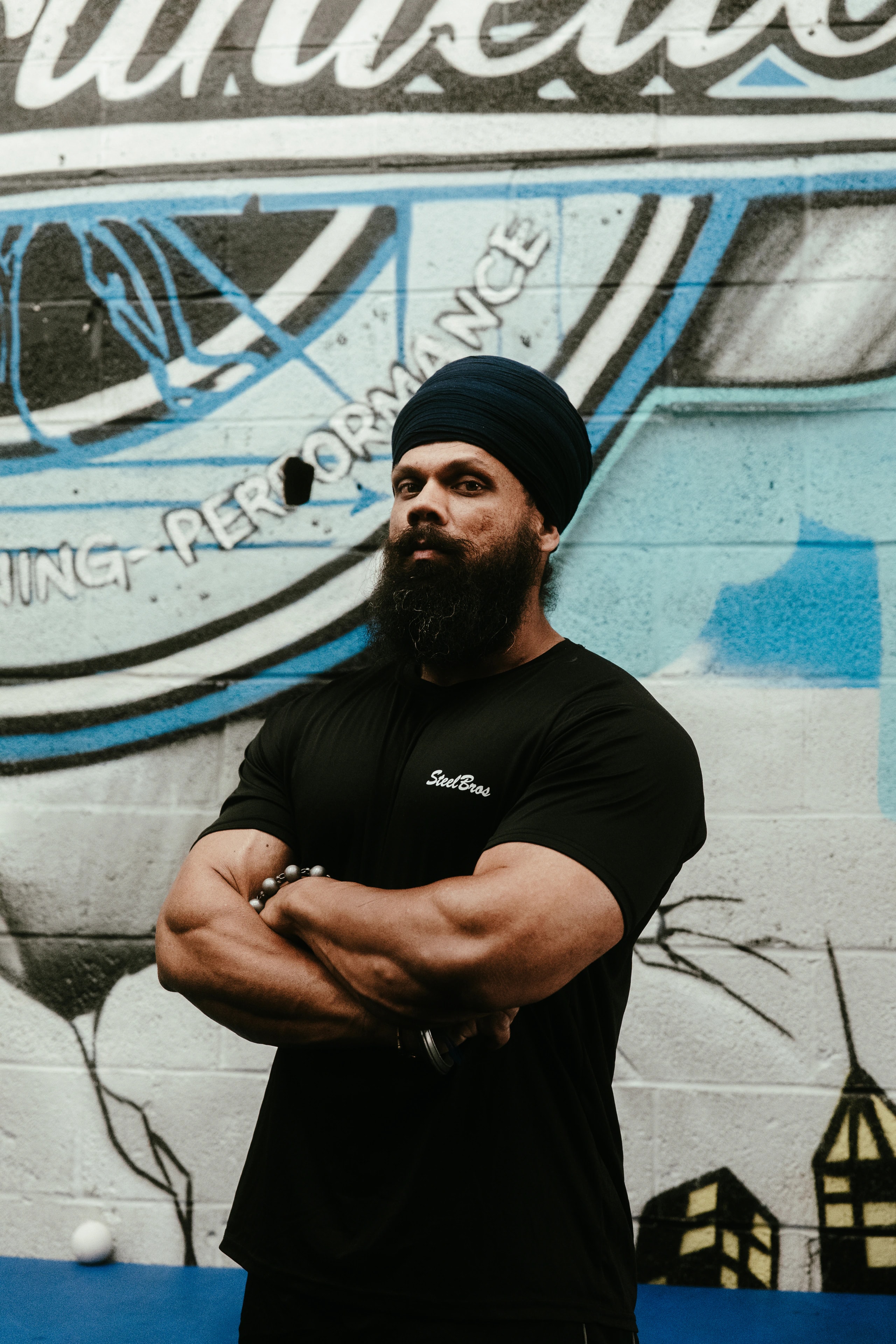 A bearded person in a black turban stands with arms crossed in front of a painted wall.
