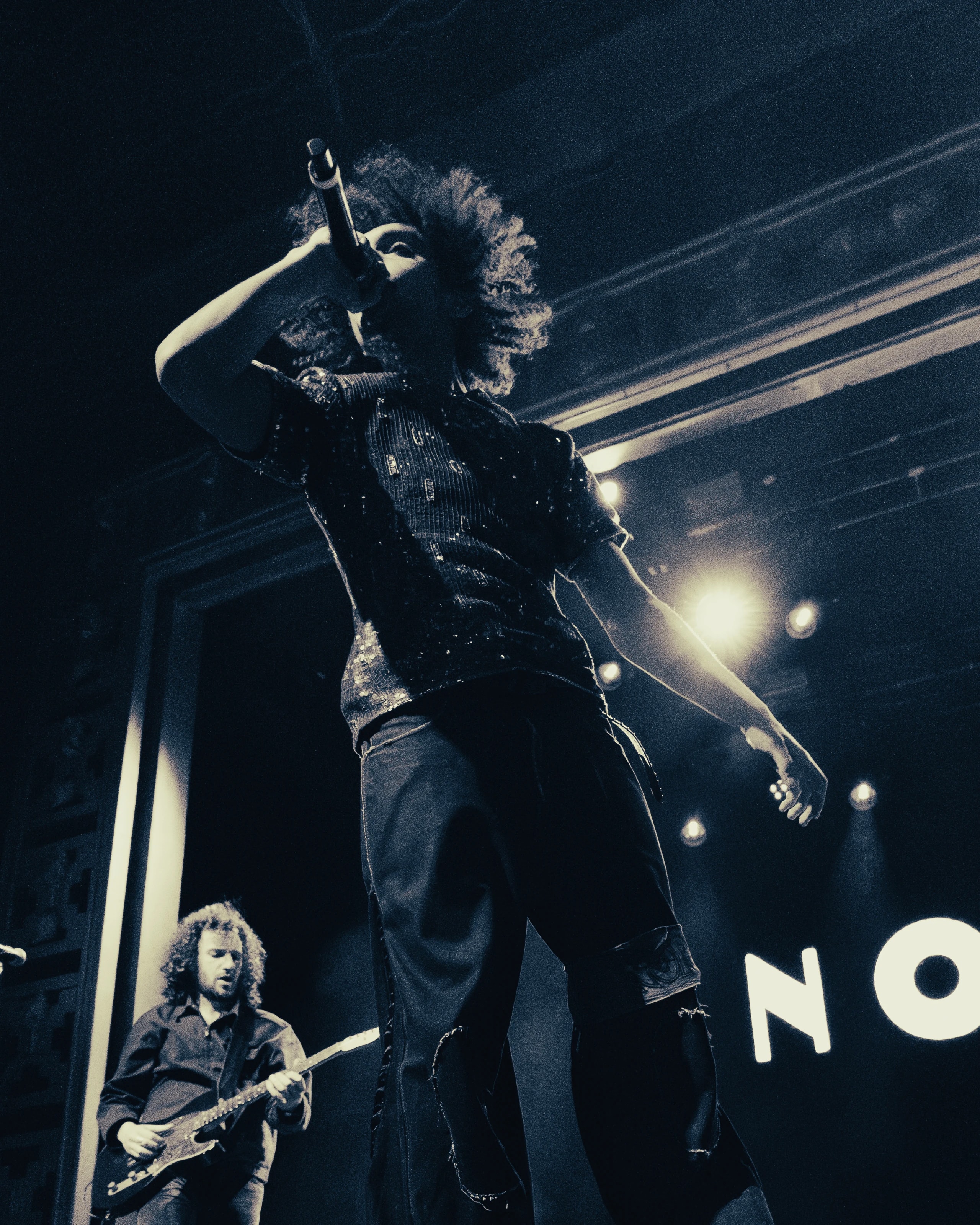 A vocalist performs from a low angle while a guitarist plays behind them under bright stage lights.
