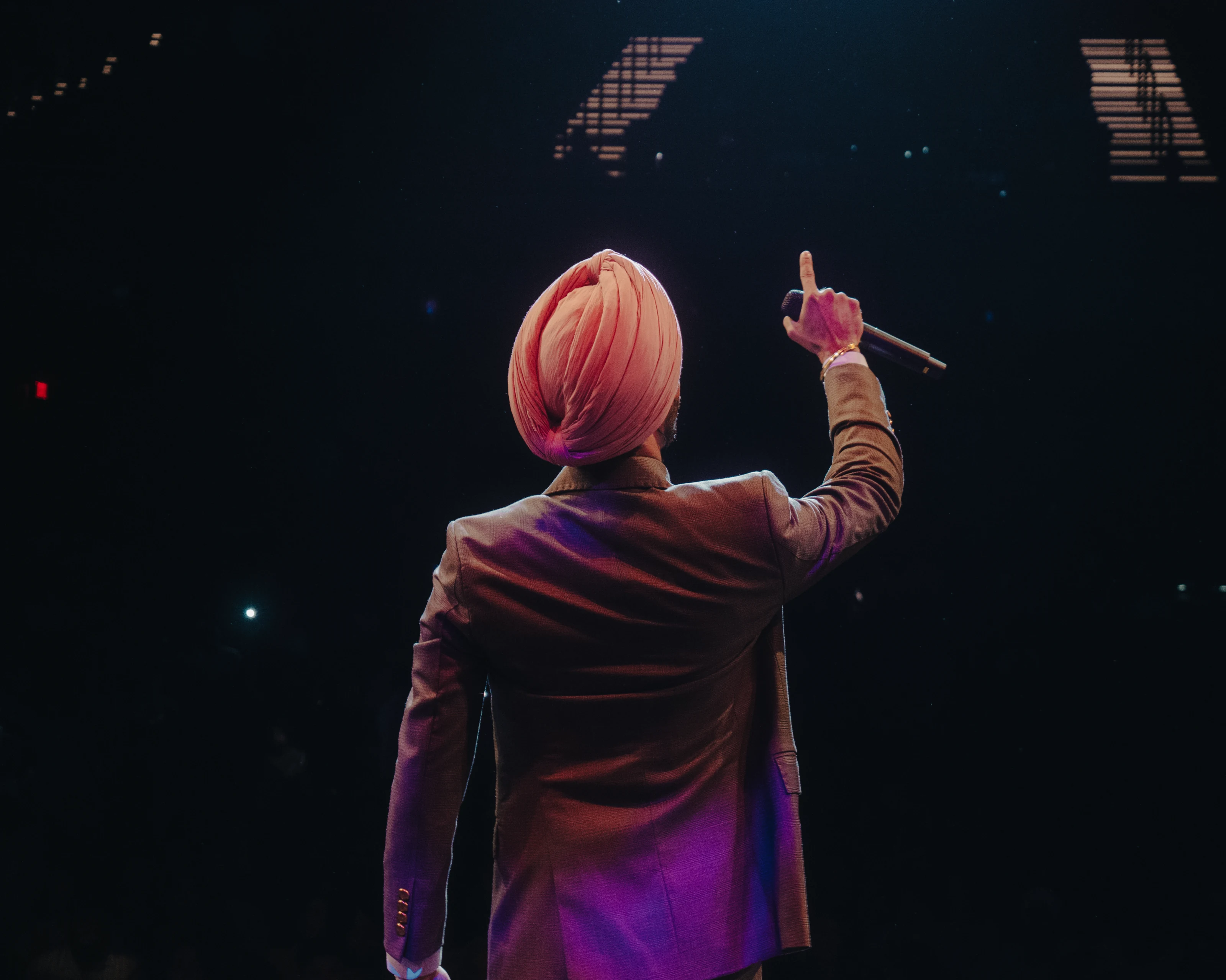 Nirvair Pannu, seen from behind in a pink turban and suit, raises a microphone toward an arena audience.