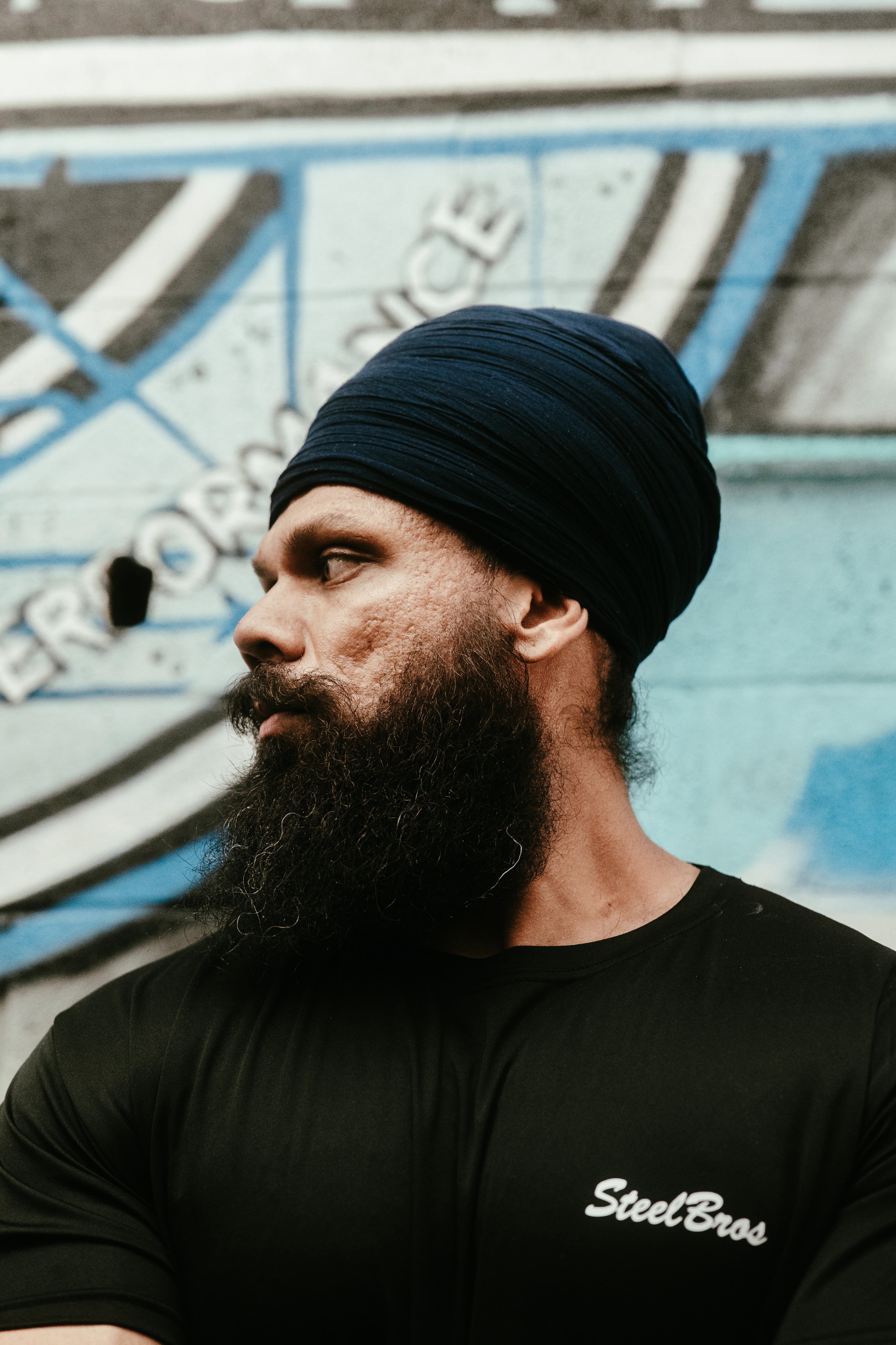 A bearded person in a black turban stands in front of a blue-and-white painted wall.
