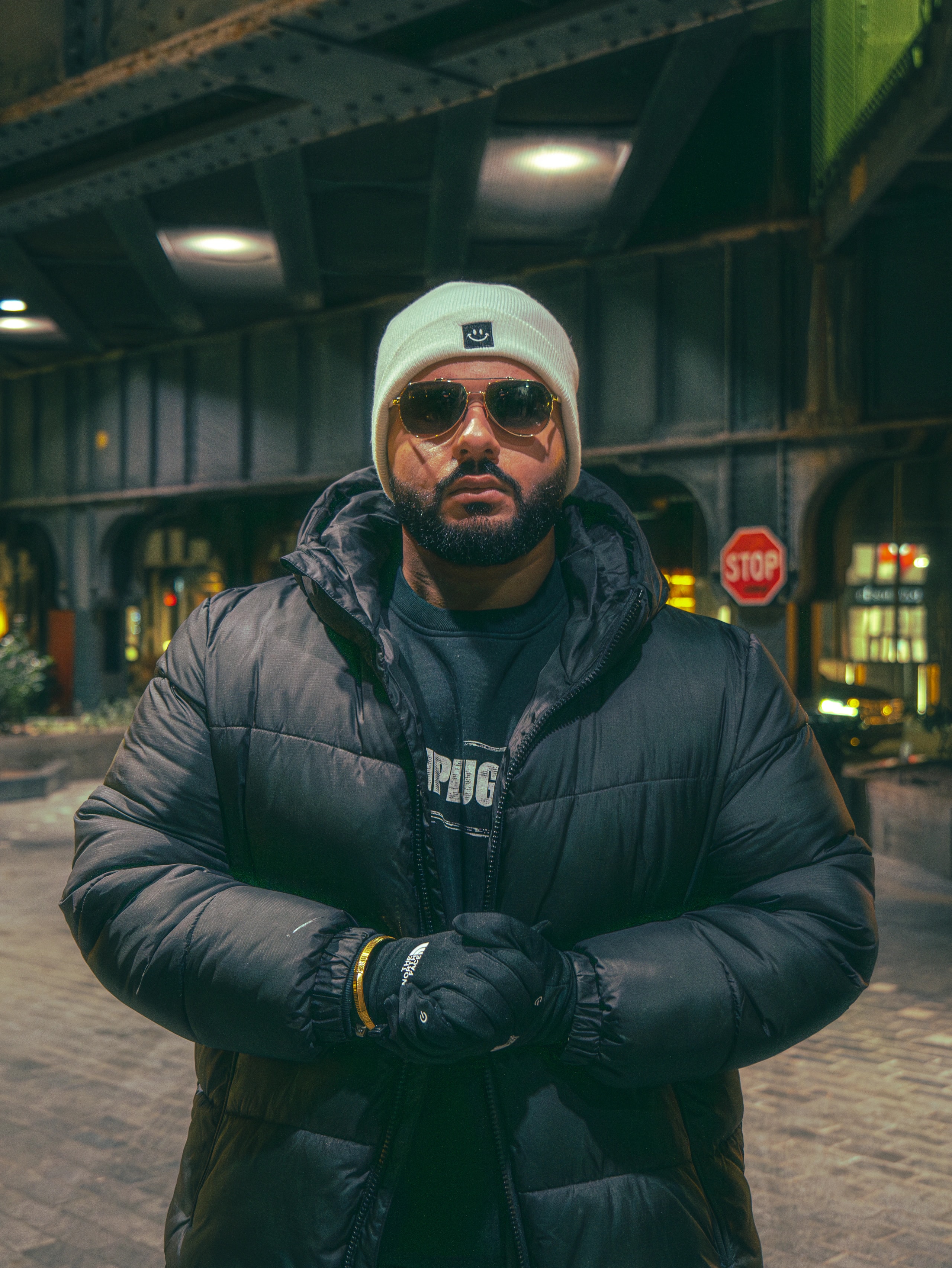A person in a white knit hat and sunglasses stands beneath an overpass.