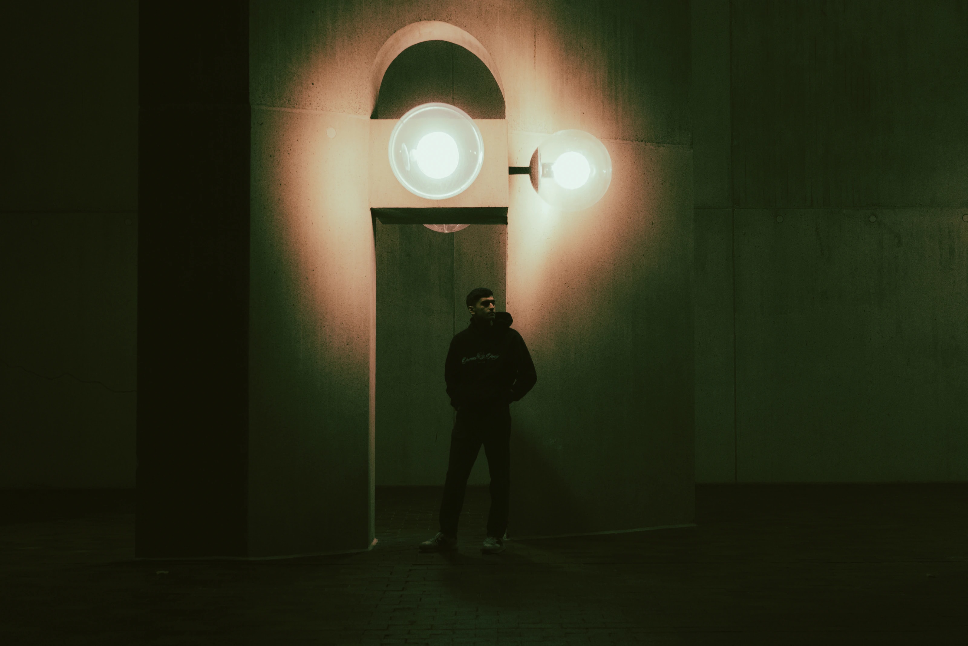 A person stands beneath two bright lamps and a rounded archway at night.